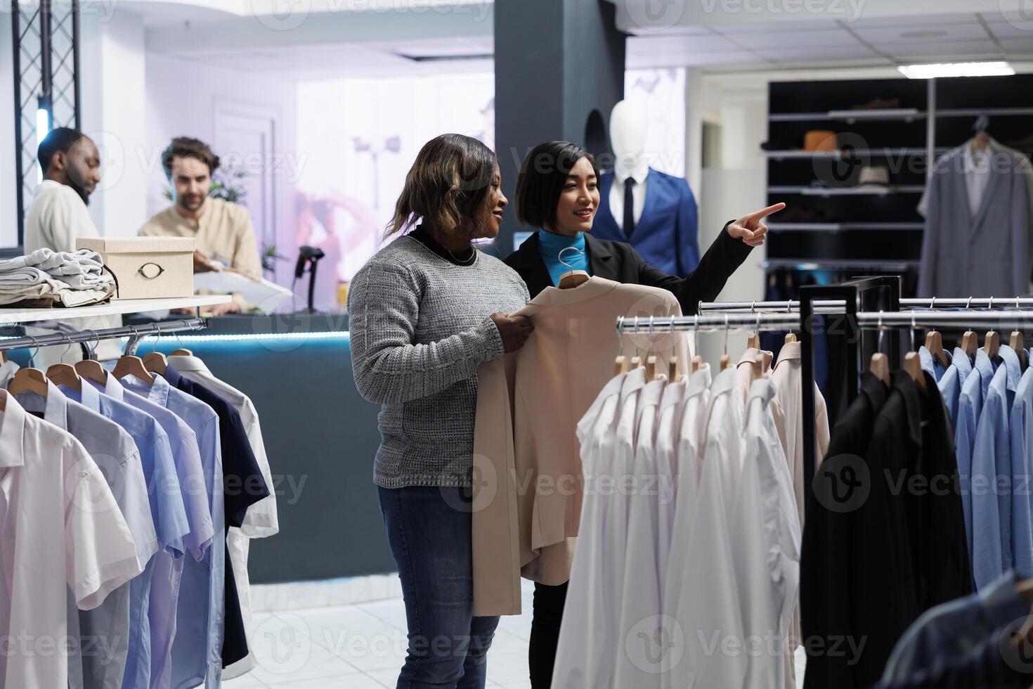 Clothing store asian woman worker pointing with finger at apparel rack while customer asking about jacket size options. Mall employee helping client to choose formal outfit and giving advice photo