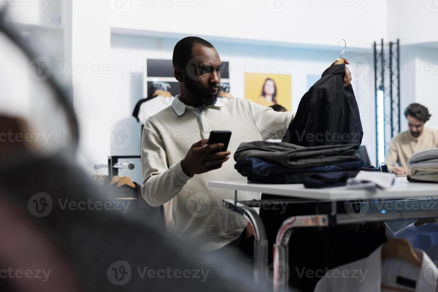 African american man choosing casual jacket in clothing store and checking price on website using mobile phone. Department mall client browsing apparel rack and examining boutique app photo