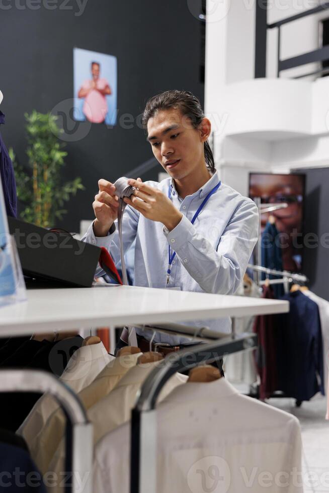 Asian man putting elegant belt on store shelf, arranging stylish accessories in modern boutique. Shopping mall employee working with fashionable clothes. Fashion new collection concept photo