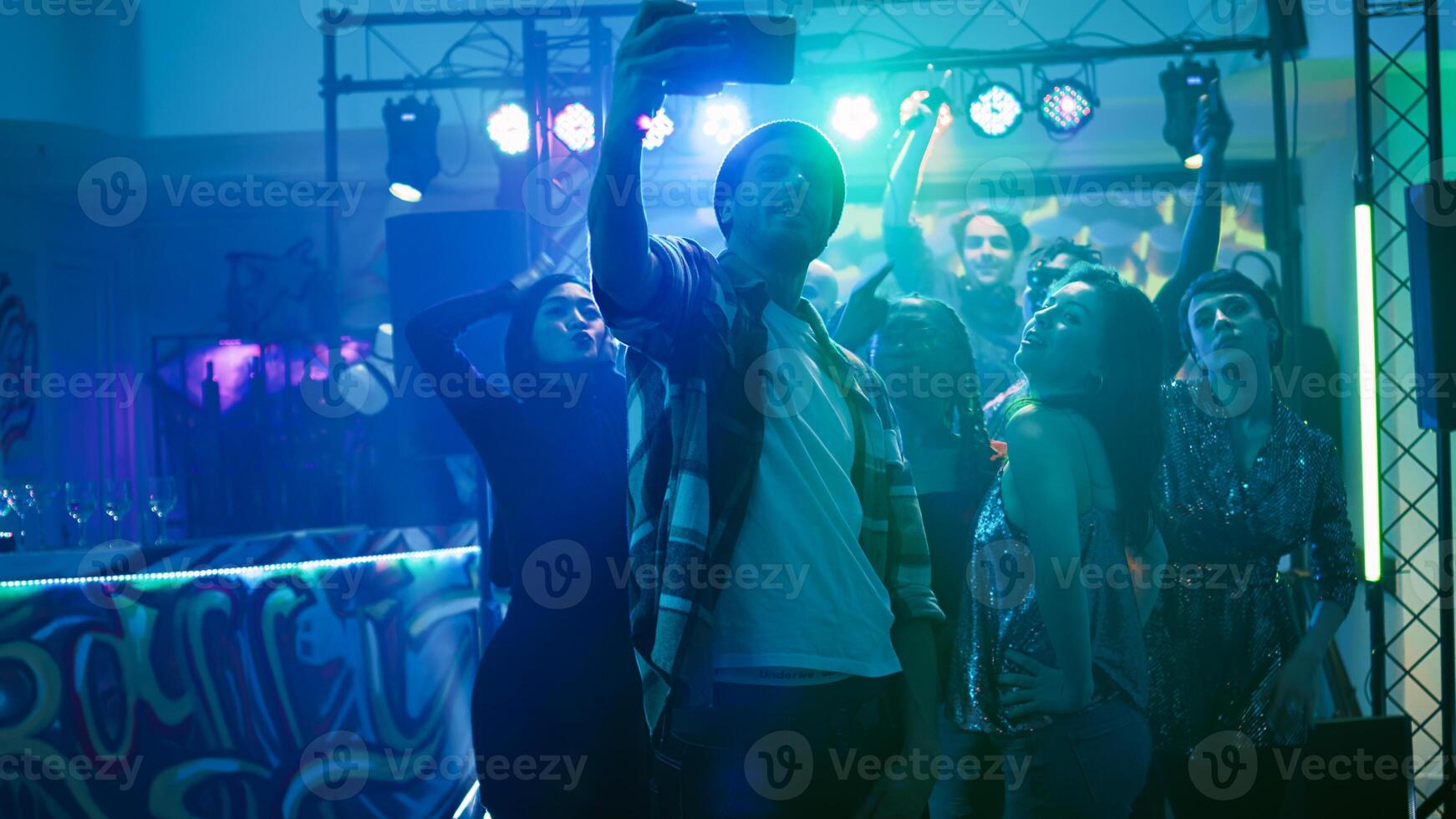 Friends taking pictures at nightclub, partying together under discolights. Group of men and women having fun at party event with electronic music, taking photos and filming. Handheld shot.