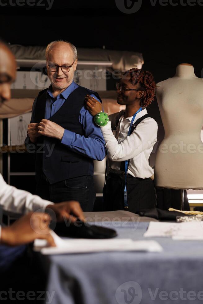 Cheerful meticulous seamstress doing precise additional needlework needed on sartorial piece worn by elderly client. Skilled african american tailor helping customer with bespoke suit comission photo