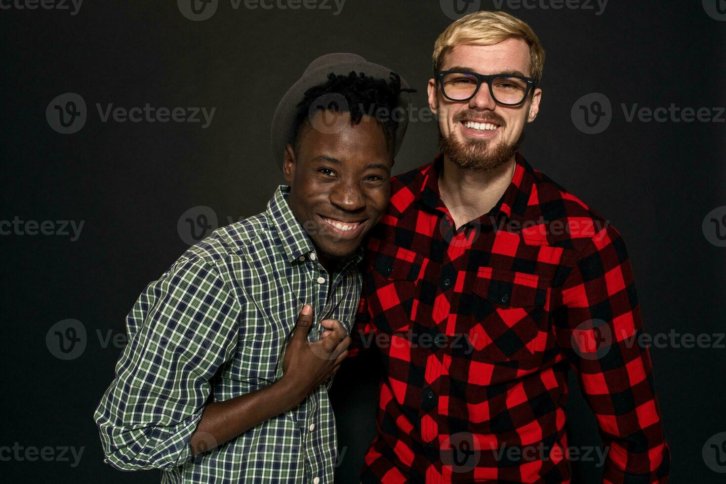 estudio Disparo de dos elegante joven hombres teniendo divertida. hermoso barbado hipster en un camisa en un jaula en pie siguiente a su afroamericano amigo en sombrero en contra un oscuro antecedentes. foto