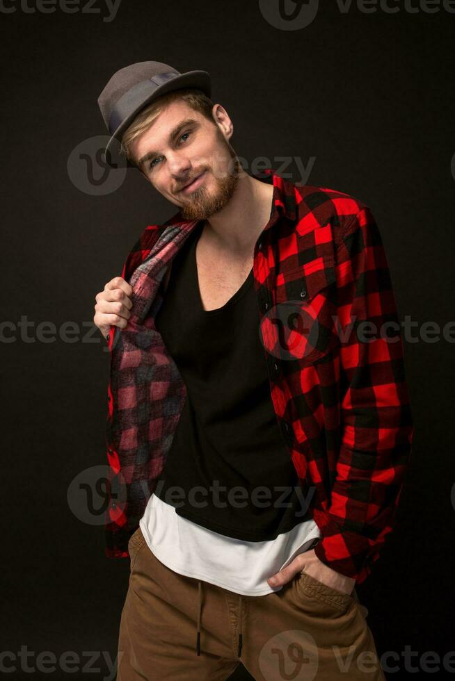 Handsome trendy young guy at the studio on black background. He wears beard and a plaid shirt photo