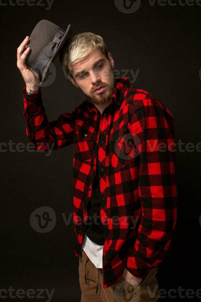 Handsome trendy young guy at the studio on black background. He wears beard and a plaid shirt photo