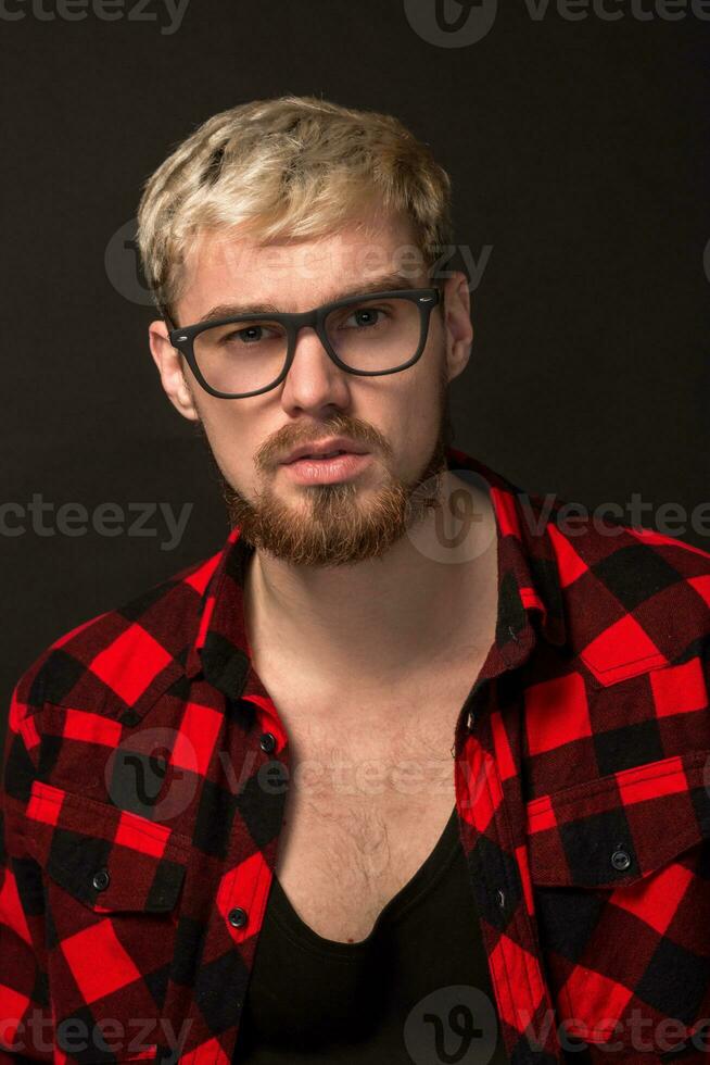 Handsome trendy young guy at the studio on black background. He wears beard and a plaid shirt photo