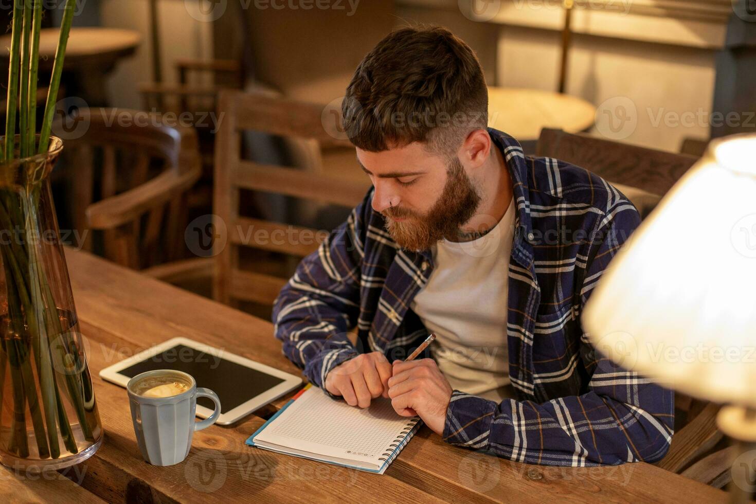 Young bearded businessman sits in cafe, home at table and writes in notebook, near lies tablet computer with black screen. Man is working, studying. photo