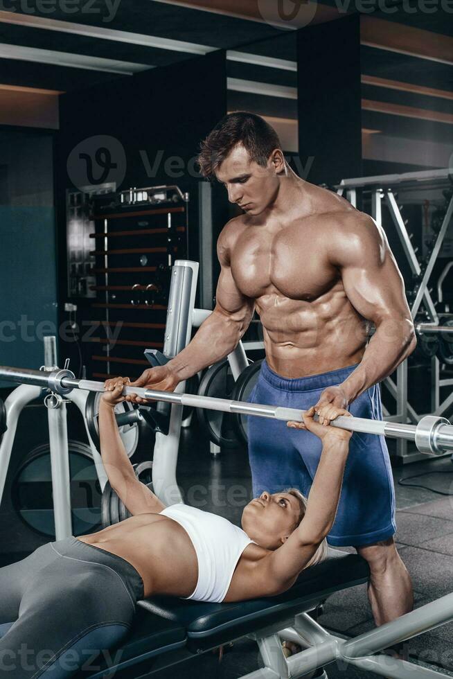 Personal trainer helping a young woman lift a barbell while working out in a gym photo