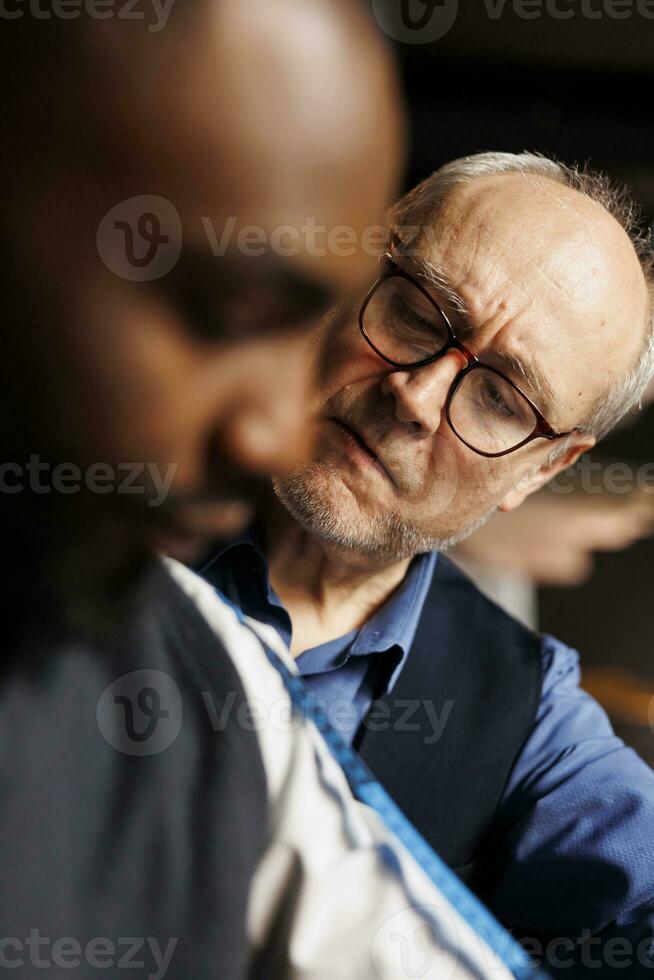 African american customer getting arm measurements taken by expert elderly tailor in atelier workspace. Experienced senior suitmaker working on handmade bespoke suit piece comission, close up photo