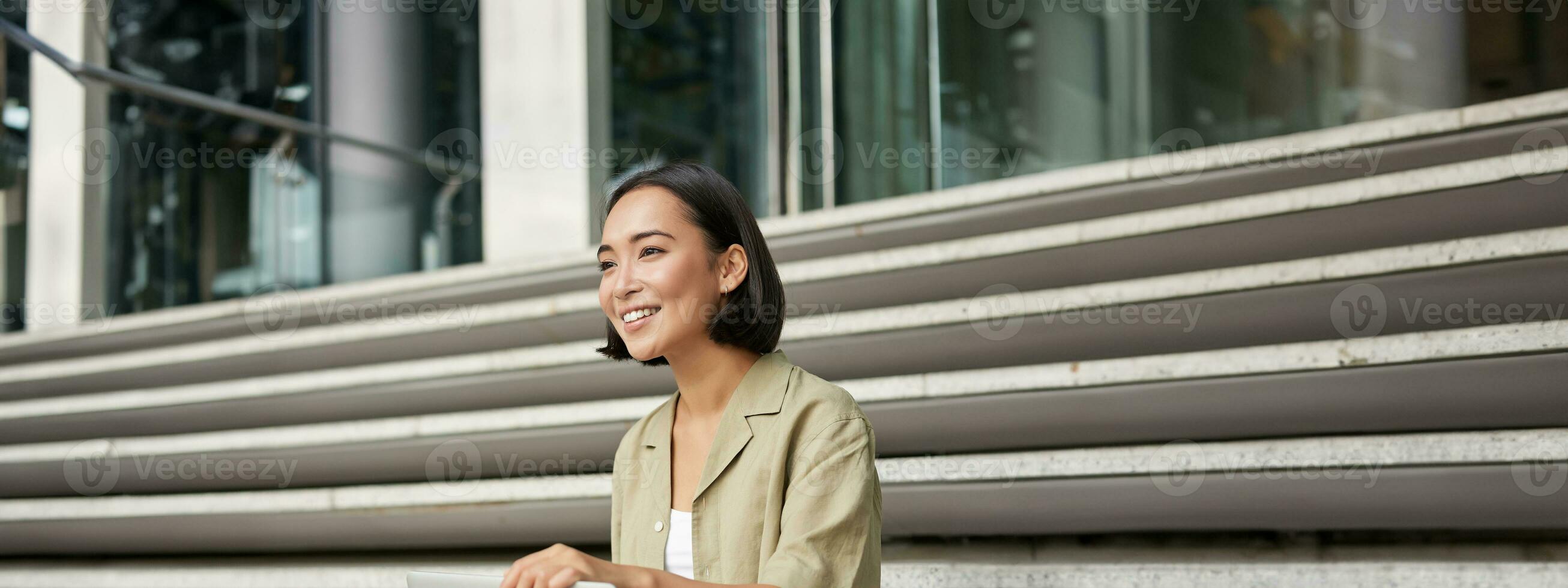 Vertical shot of smiling asian woman with laptop computer, sitting on stairs outside building. photo