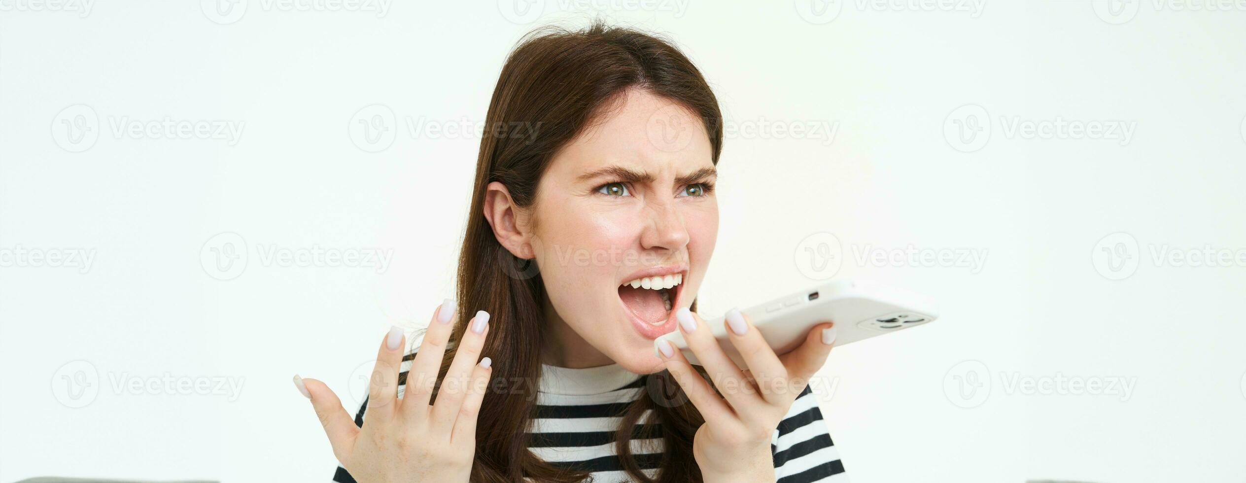 Portrait of angry woman shouting at smartphone speakerphone, recording voice message with annoyed face expression, screaming at phone, white background photo