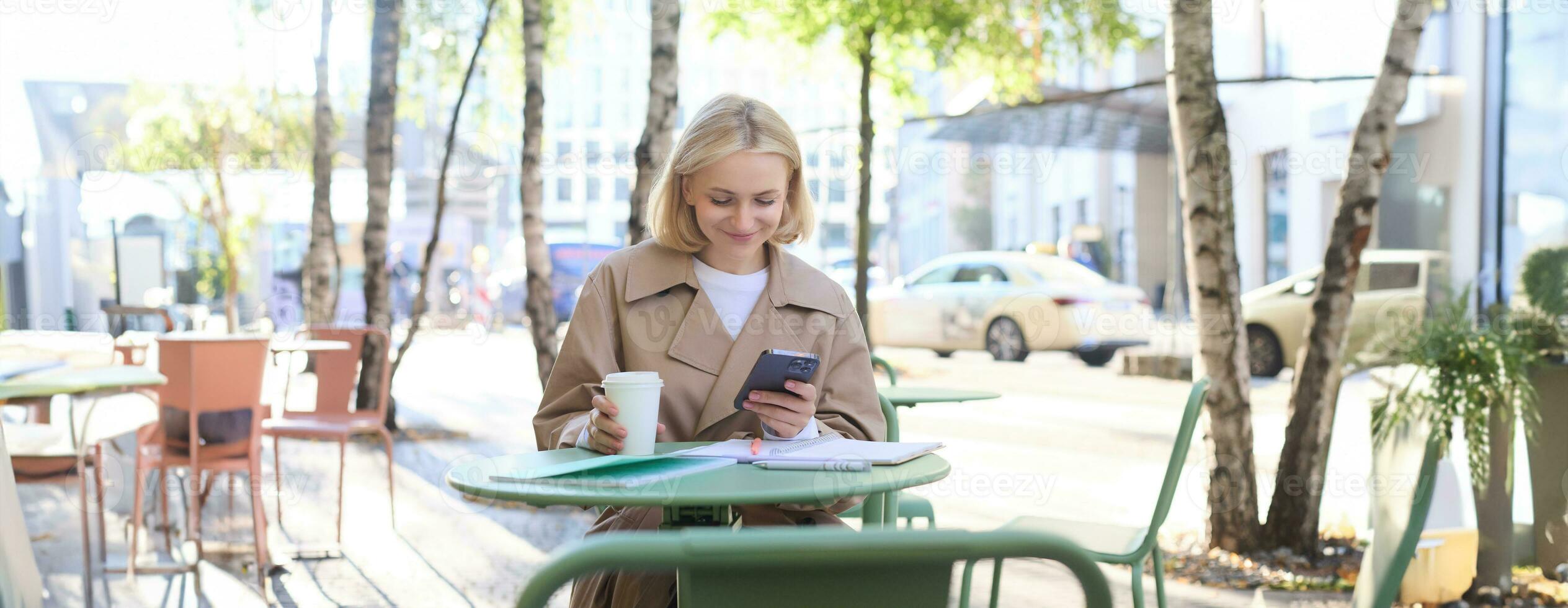 Image of young woman drinking coffee, sitting in outdoor cafe with smartphone, sending message, reading notification on mobile phone and smiling photo