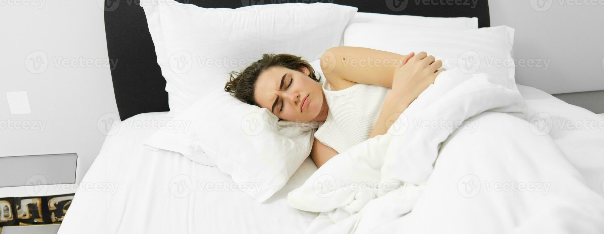 Portrait of woman feeling unwell, lying in bed and touching her belly, frowning from painful menstrual cramps, having period pain, resting in bedroom photo