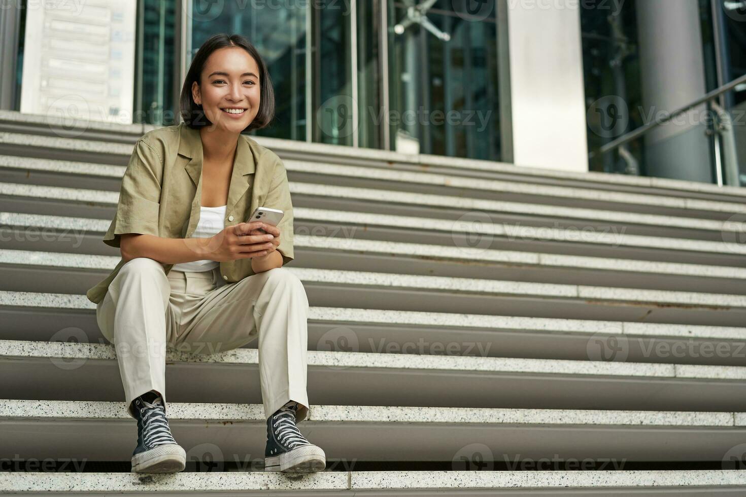 Portrait of smiling asian girl sits on stairs outdoors, sending message, using smartphone app, looking at mobile phone screen photo