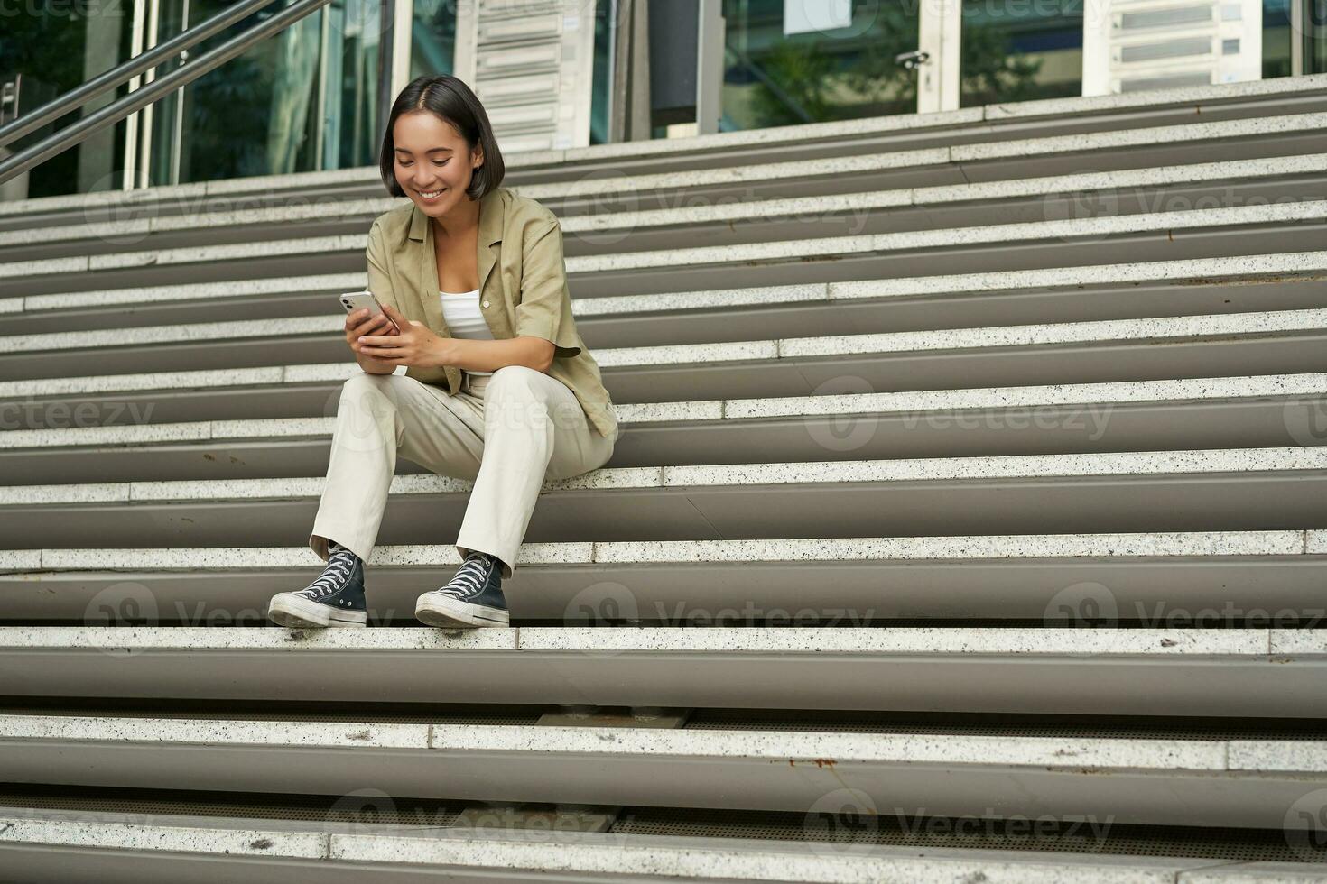 Portrait of smiling asian girl sits on stairs outdoors, sending message, using smartphone app, looking at mobile phone screen photo