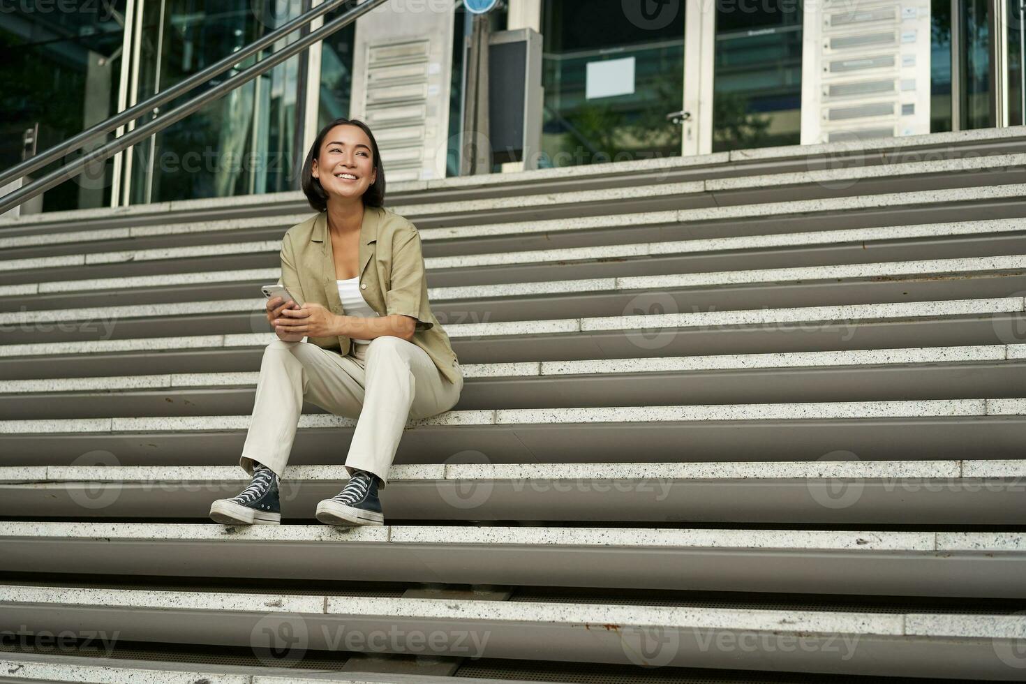 Portrait of smiling asian girl sits on stairs outdoors, sending message, using smartphone app, looking at mobile phone screen photo