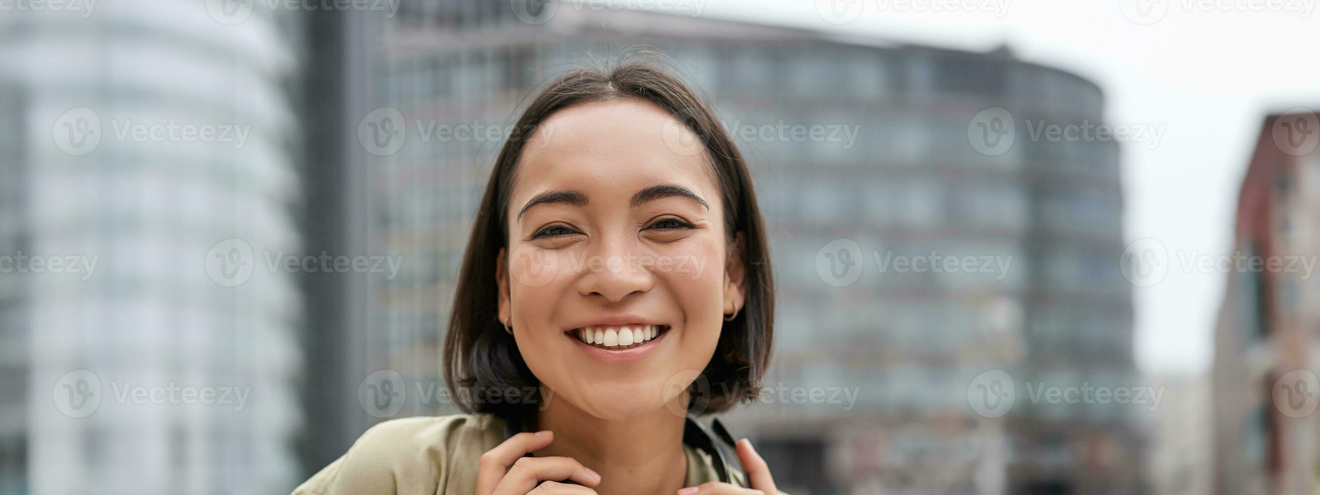 Vertical shot of beautiful asian woman posing with headphones around neck, smiling and laughing, standing on street in daylight photo