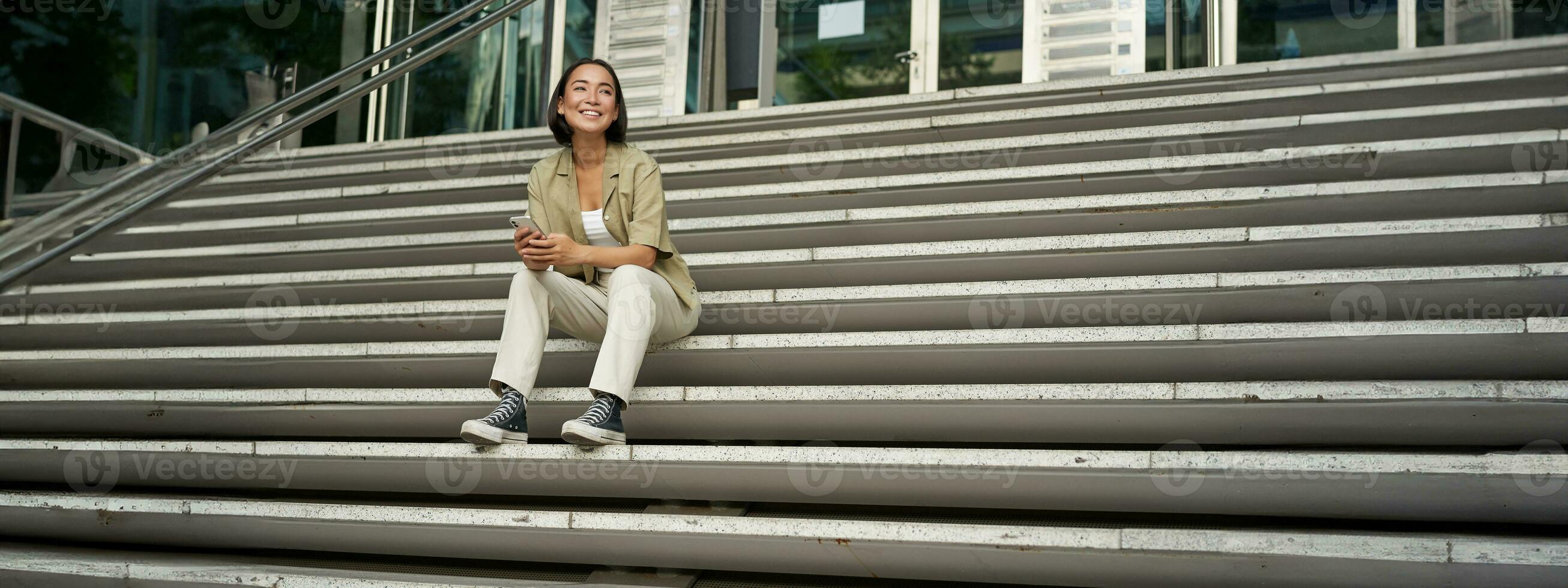 Portrait of smiling asian girl sits on stairs outdoors, sending message, using smartphone app, looking at mobile phone screen photo