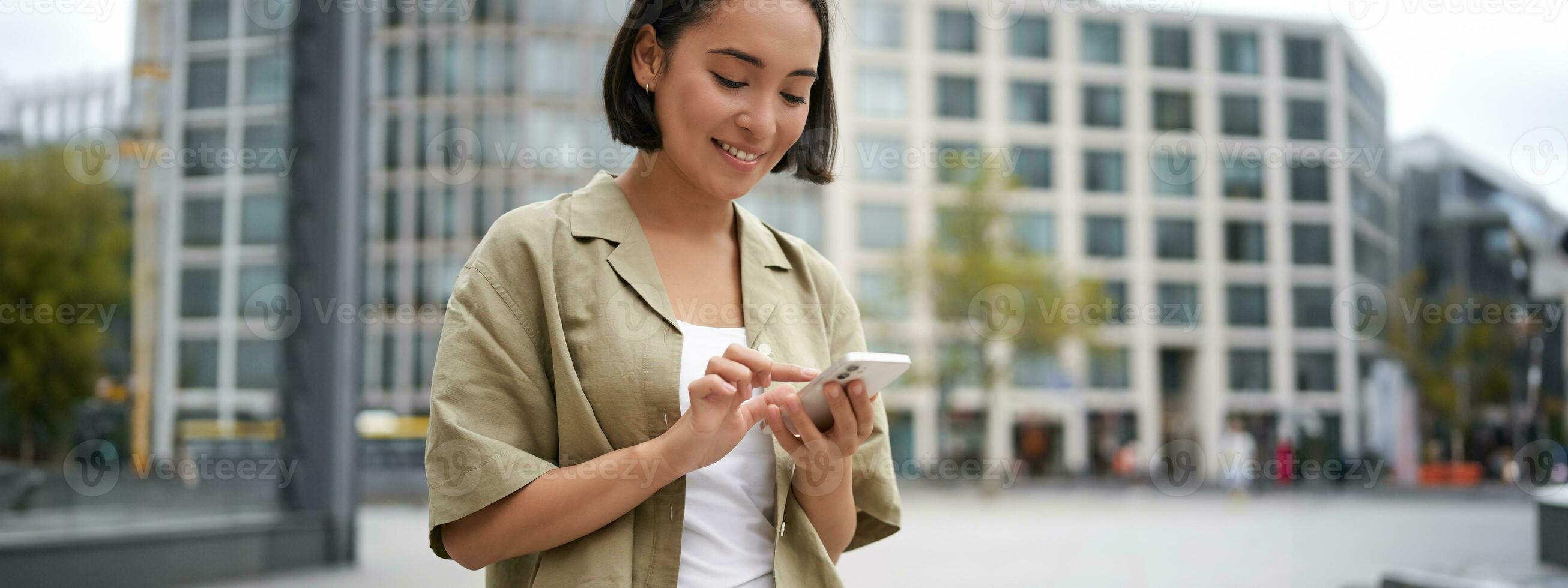 Mobile technology. Smiling asian woman using smartphone app, looking at her telephone on street, checking map, calling or texting someone photo
