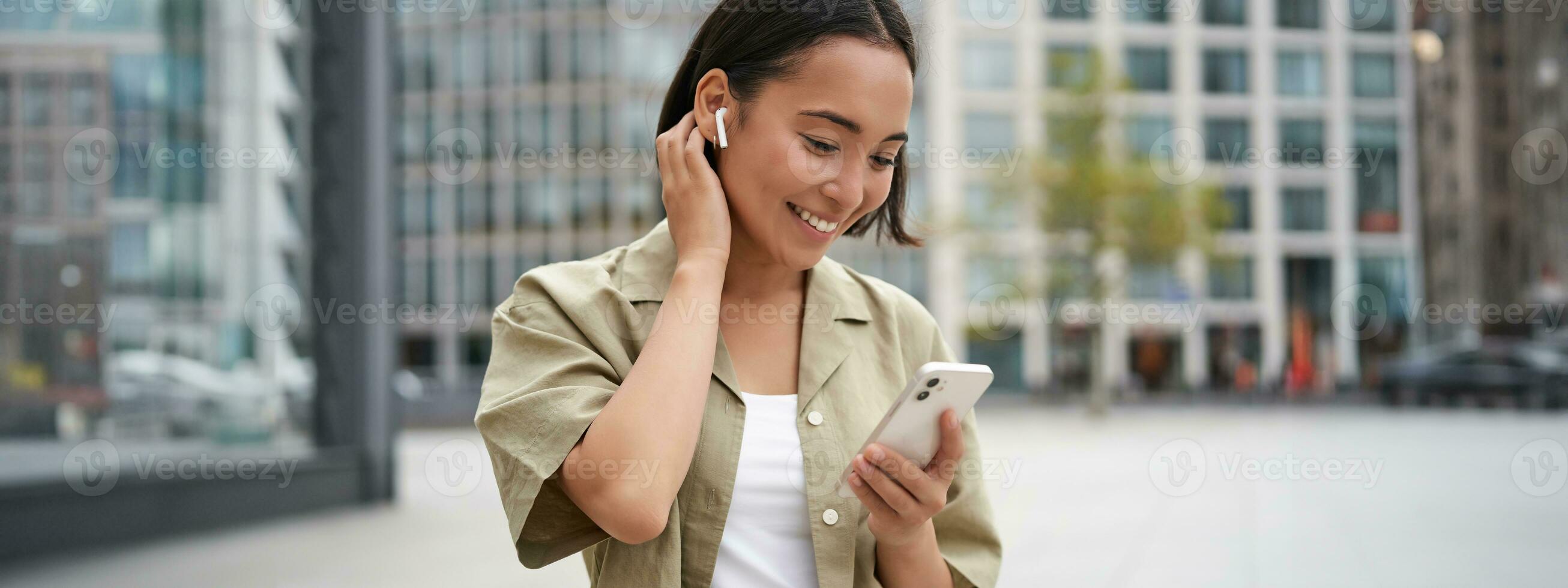 Smiling asian girl listens music in wireless headphones, looks at her phone, choosing music or podcast. Young woman calling someone, using headphones photo