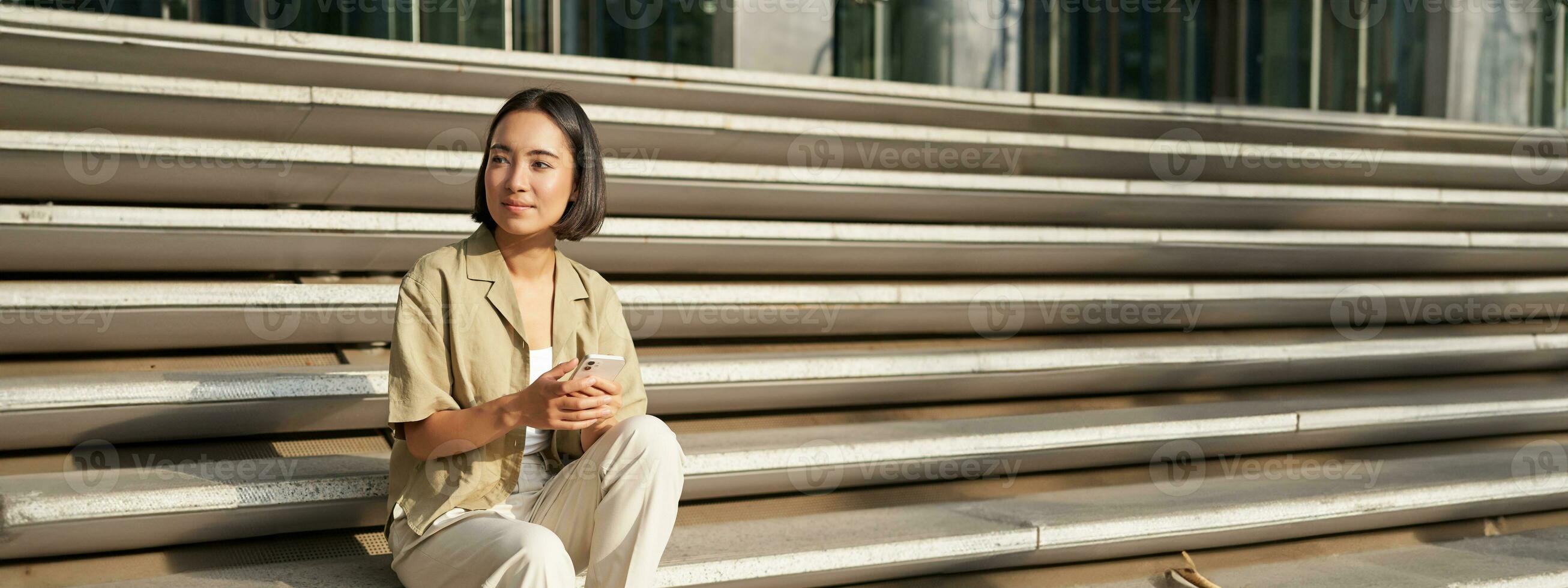 Beautiful asian girl sitting on stairs outside building, using mobile phone, looking at smartphone app and smiling photo