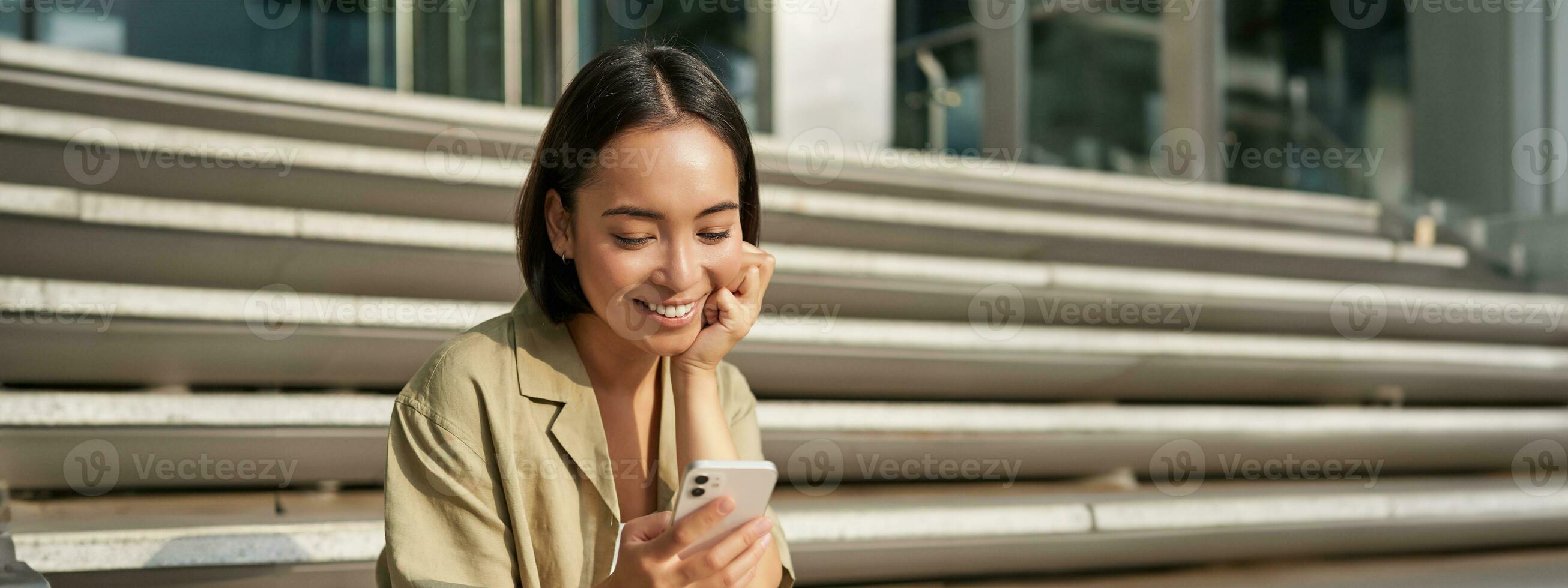 Vertical shot of asian woman, student sits on stairs in city, looking at mobile phone screen and smiling, using smartphone app photo