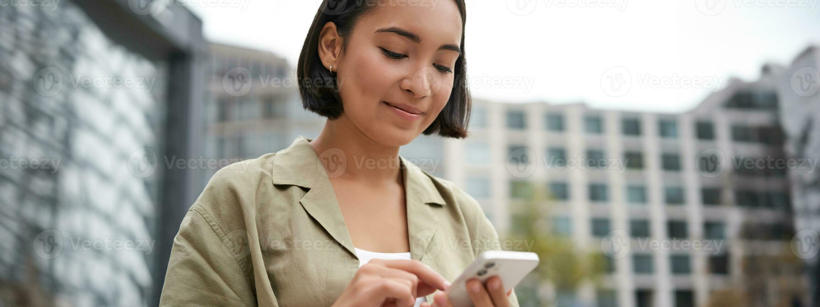 Mobile technology. Smiling asian woman using smartphone app, looking at her telephone on street, checking map, calling or texting someone photo