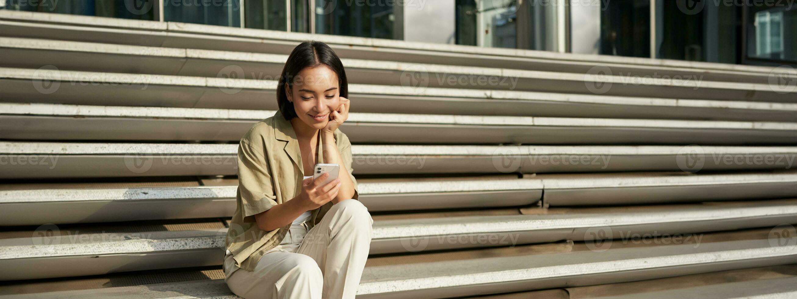 Vertical shot of asian woman, student sits on stairs in city, looking at mobile phone screen and smiling, using smartphone app photo