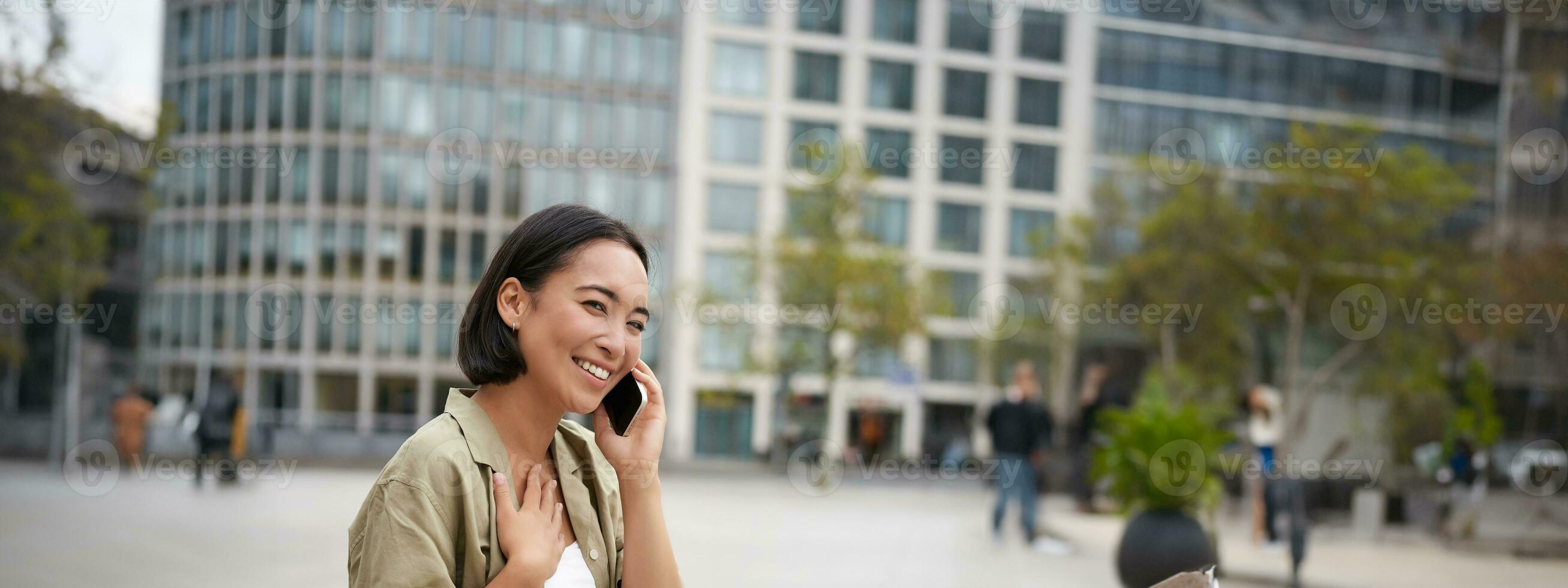 Vertical shot of smiling stylish asian girl, having a telephone conversation and walking on street. Young woman talking on mobile phone photo