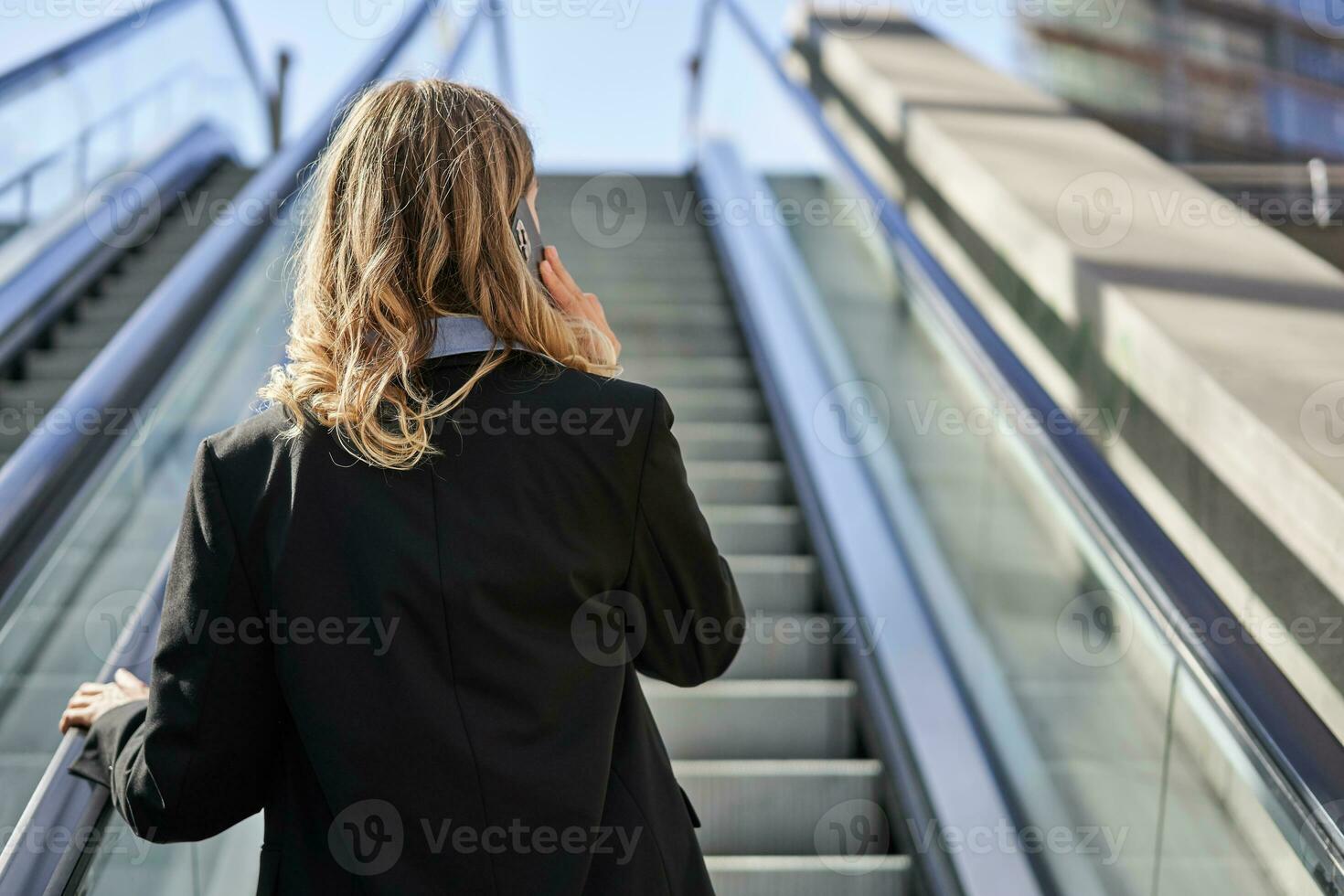 Rear view of businesswoman in suit, standing on escalator and talking on mobile phone photo