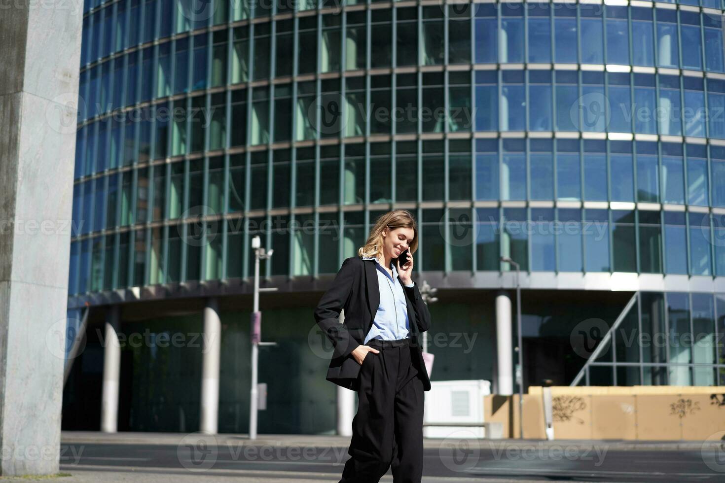 Young corporate woman in suit walking on street and talking on mobile phone. Businesswoman having telephone conversation while going to work photo