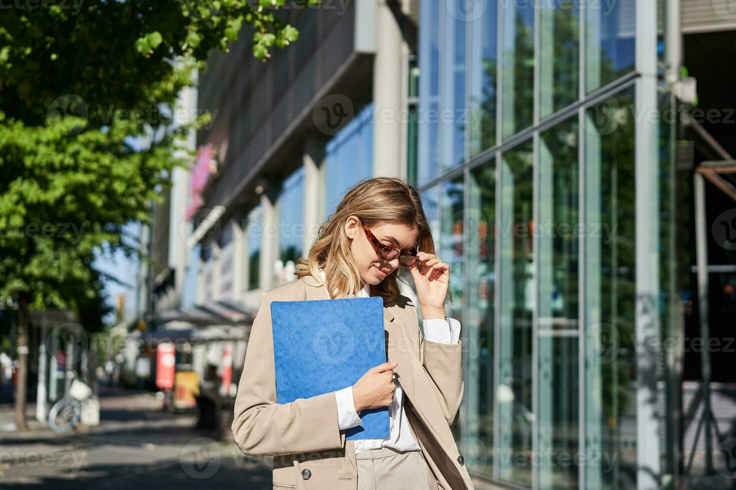 retrato de confidente corporativo mujer yendo a oficina con un carpeta, caminando en calle en soleado día en traje y Gafas de sol foto