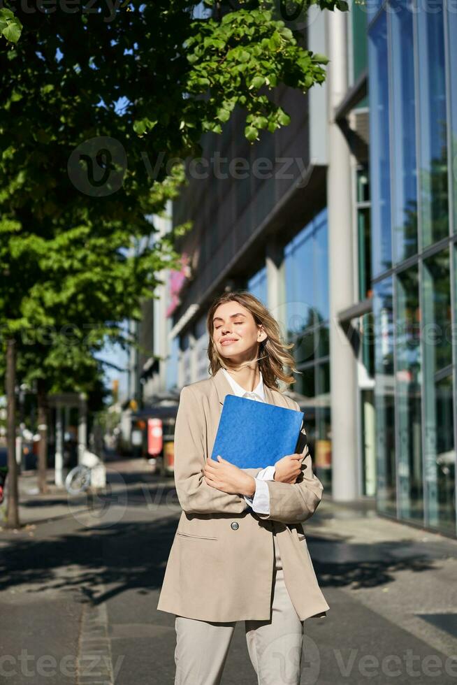 Vertical shot of businesswoman with folder, standing on a street near office buildings, wearing suit for office work photo