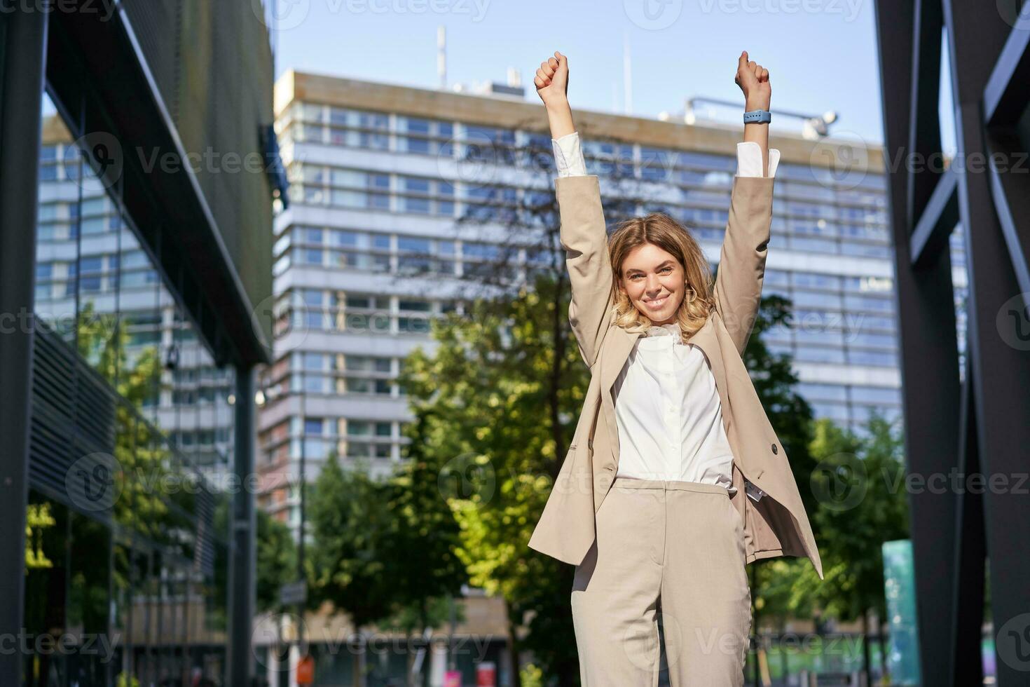 Portrait of happy young businesswoman lift hands up, does fist pump, celebrates victory, wins smth, achieves goal, stands outdoors on street photo