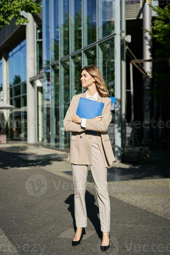 Vertical shot of businesswoman with folder, standing on a street near office buildings, wearing suit for office work photo