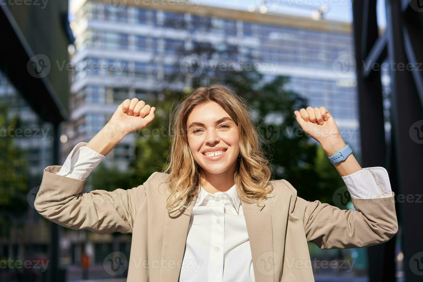 contento sonriente vendedora en beige traje, celebra su logro, triunfando, haciendo puño bomba y mirando emocionado en calle de negocio centrar foto