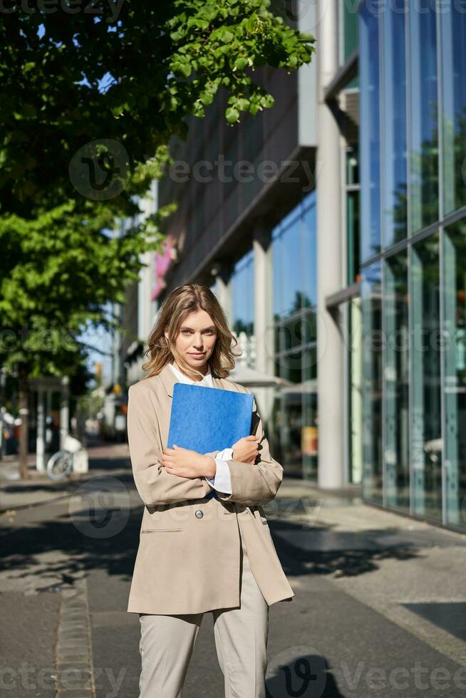 Vertical shot of businesswoman with folder, standing on a street near office buildings, wearing suit for office work photo