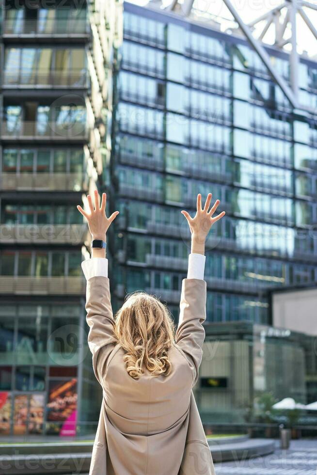 Rear view of corporate woman, lawyer celebrating, lifting hands up and triumphing, achieve goal or success, standing outside on street of city center photo