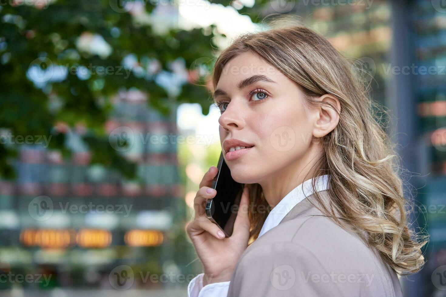 Close up shot of businesswoman talking on mobile phone. Corporate woman calling someone, looking around, standing outdoors photo
