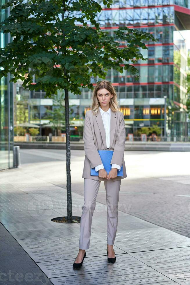 Portrait of young saleswoman in beige business suit, holding blue folder with work documents, standing outdoors on street of city center photo