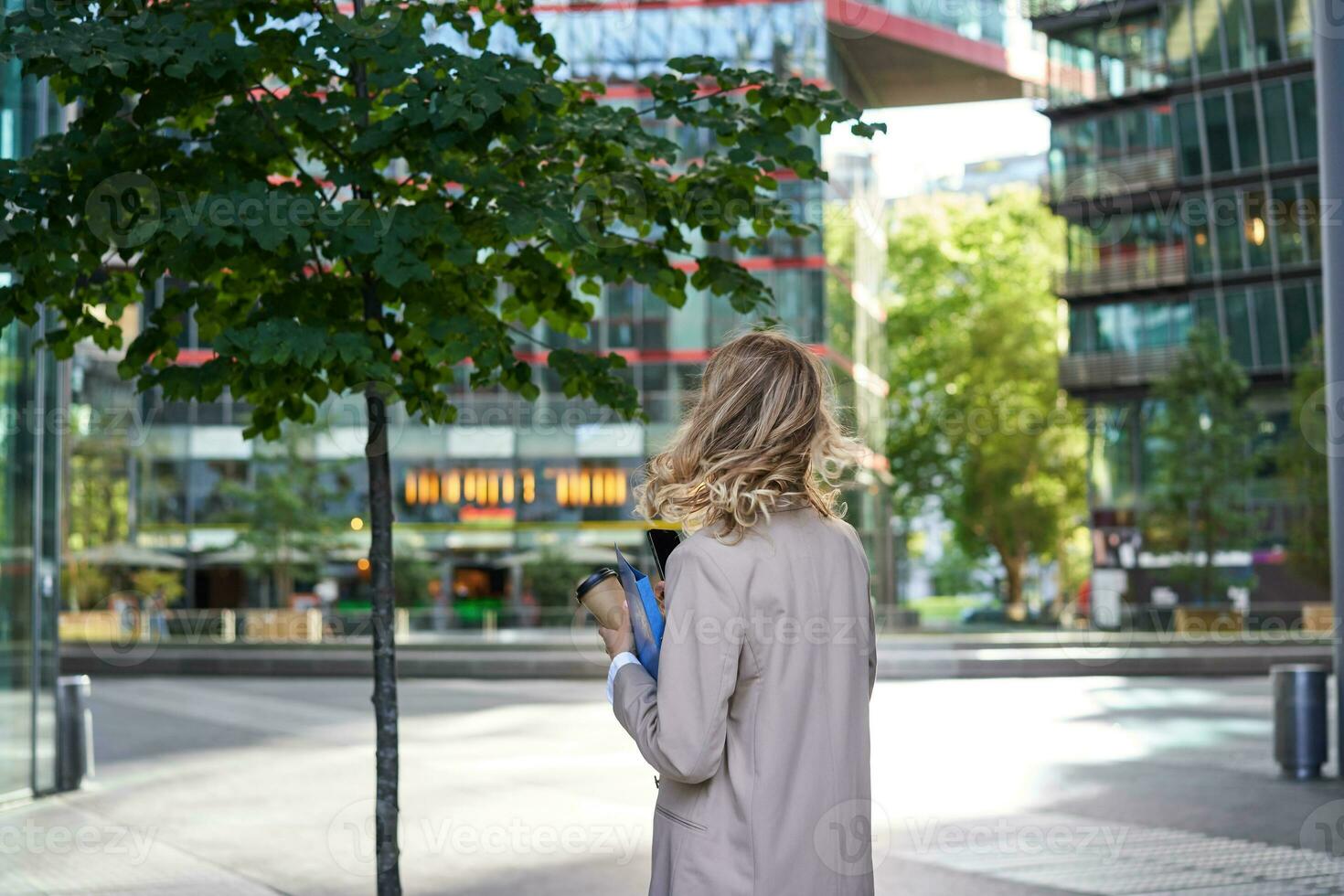 Silhouette of young business woman in beige suit, walking in city center, holding working documents and papers, standing outdoors near office buildings photo