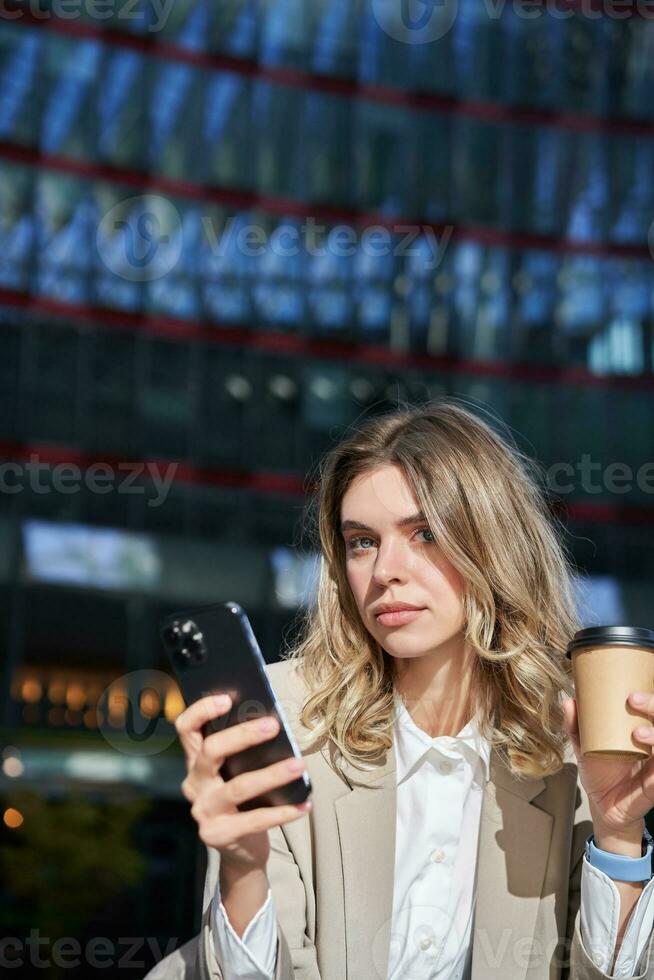 Stylish young corporate woman standing in street, drinks coffee and holds mobile phone photo