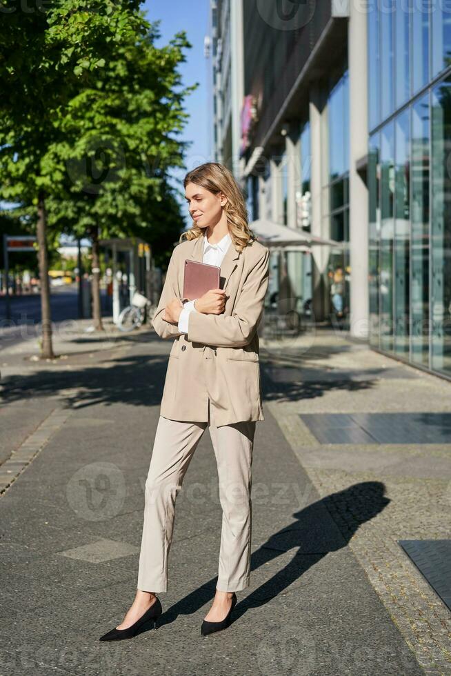 Vertical shot of businesswoman walking on street with digital tablet, going to work, wearing beige suit and high heels photo