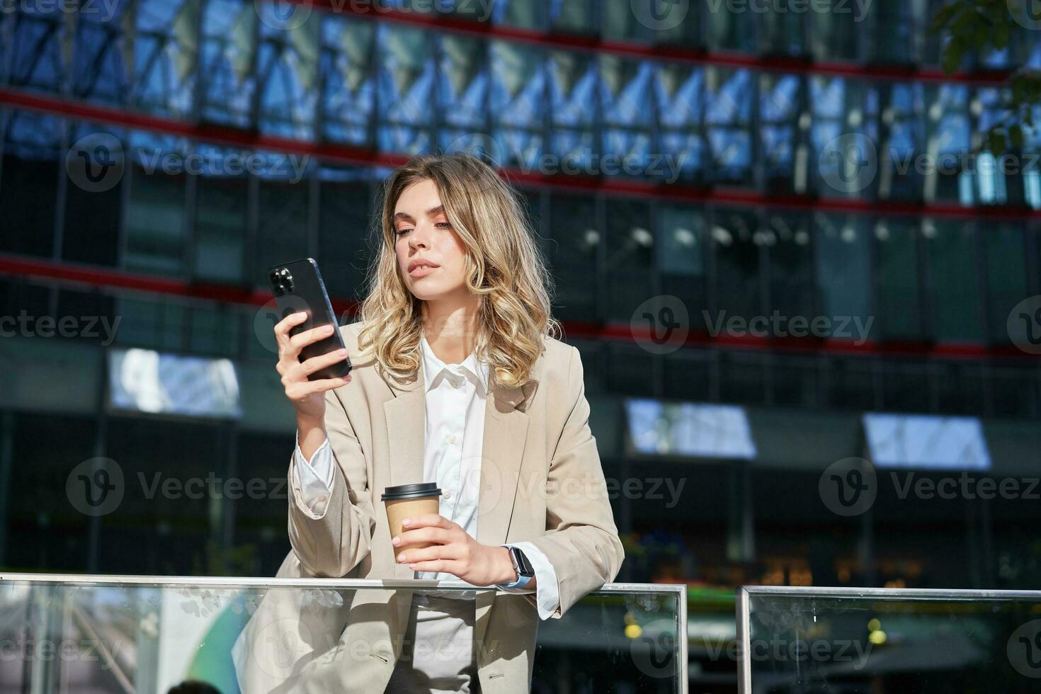 Businesswoman in suit standing with smartphone on street, drinking coffee, using mobile phone app, browsing internet, having day break photo