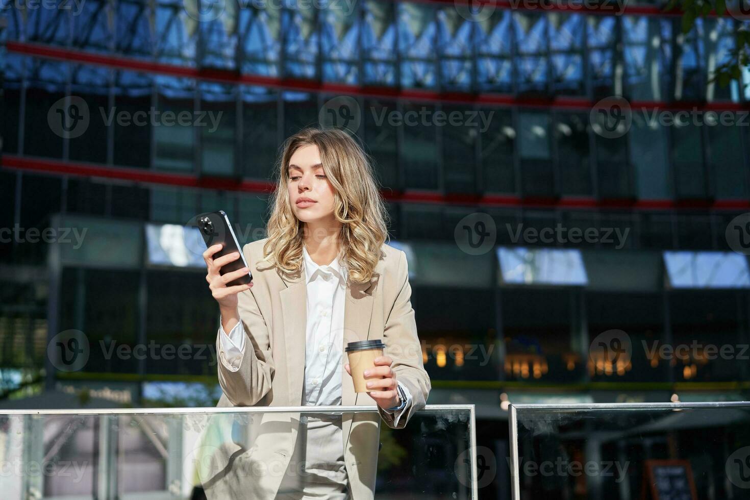 Businesswoman in suit standing with smartphone on street, drinking coffee, using mobile phone app, browsing internet, having day break photo