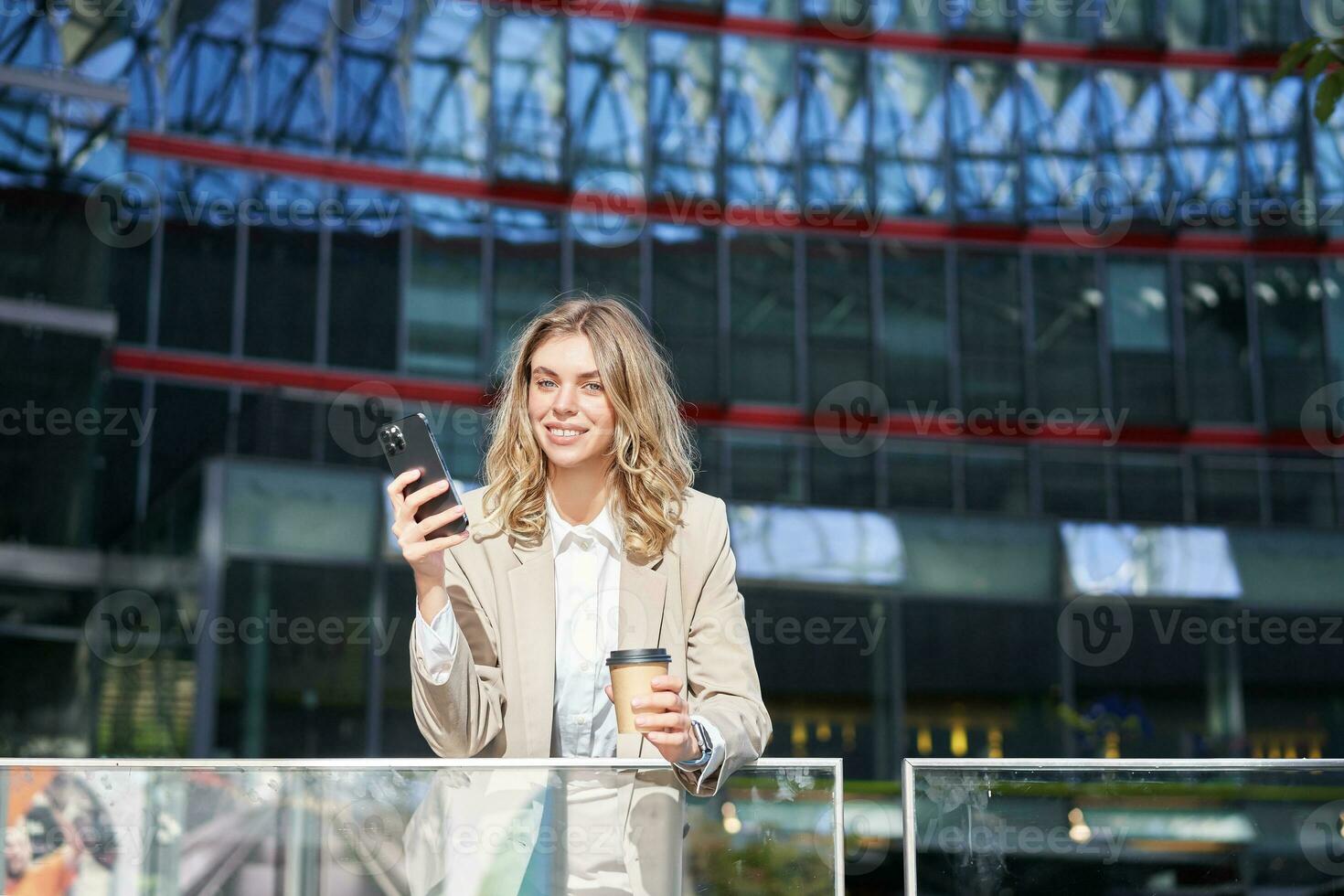 Businesswoman in suit standing with smartphone on street, drinking coffee, using mobile phone app, browsing internet, having day break photo