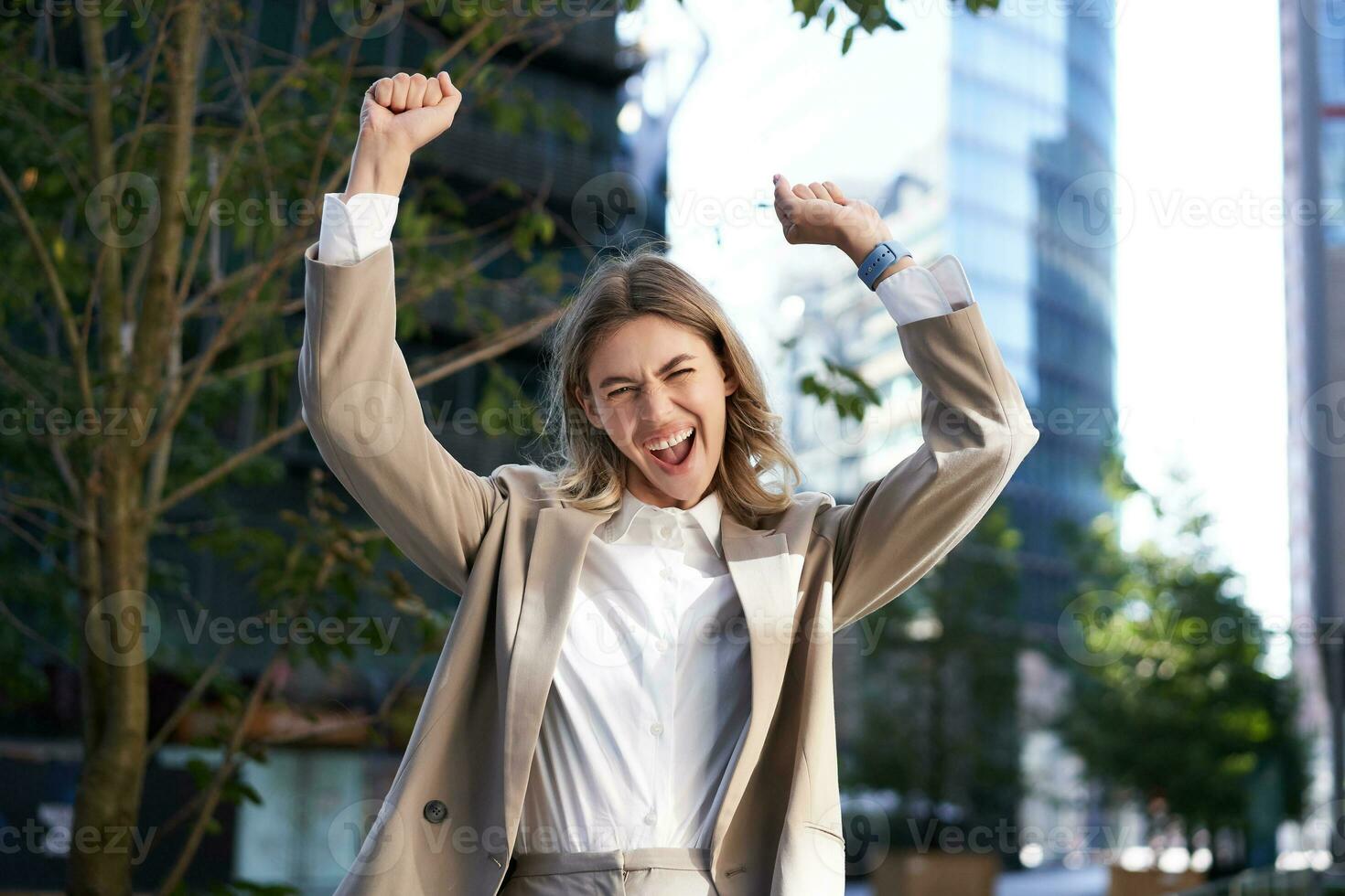 Excited businesswoman screams and celebrates, lifts hands up, does hooray gesture, celebrates victory, achieve goal, stands on street photo