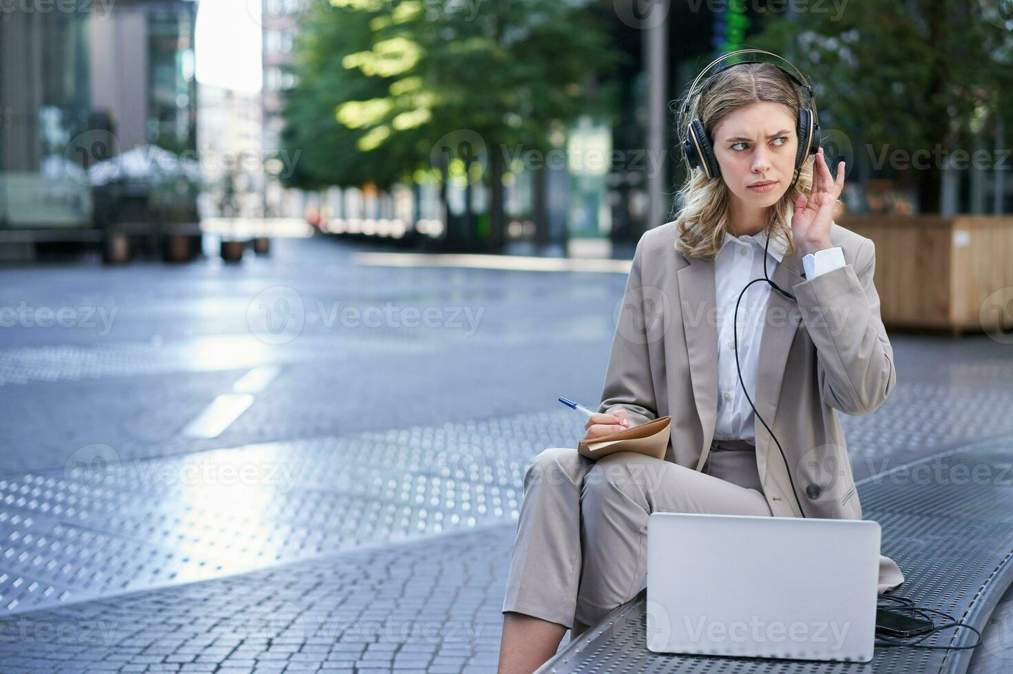 mujer sentado en un calle con ordenador portátil y auriculares atascado en, tomando notas corporativo trabajador atender en línea equipo reunión y escritura abajo información, trabajando al aire libre foto