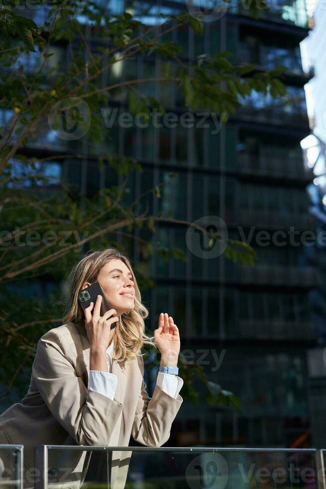 Vertical shot of businesswoman on a call, standing outside business center, talking on mobile phone photo