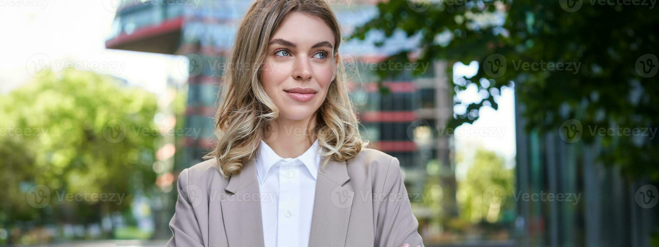 Portrait of young successful businesswoman in suit, cross arms on chest, smile and look confident, standing on street of business center photo