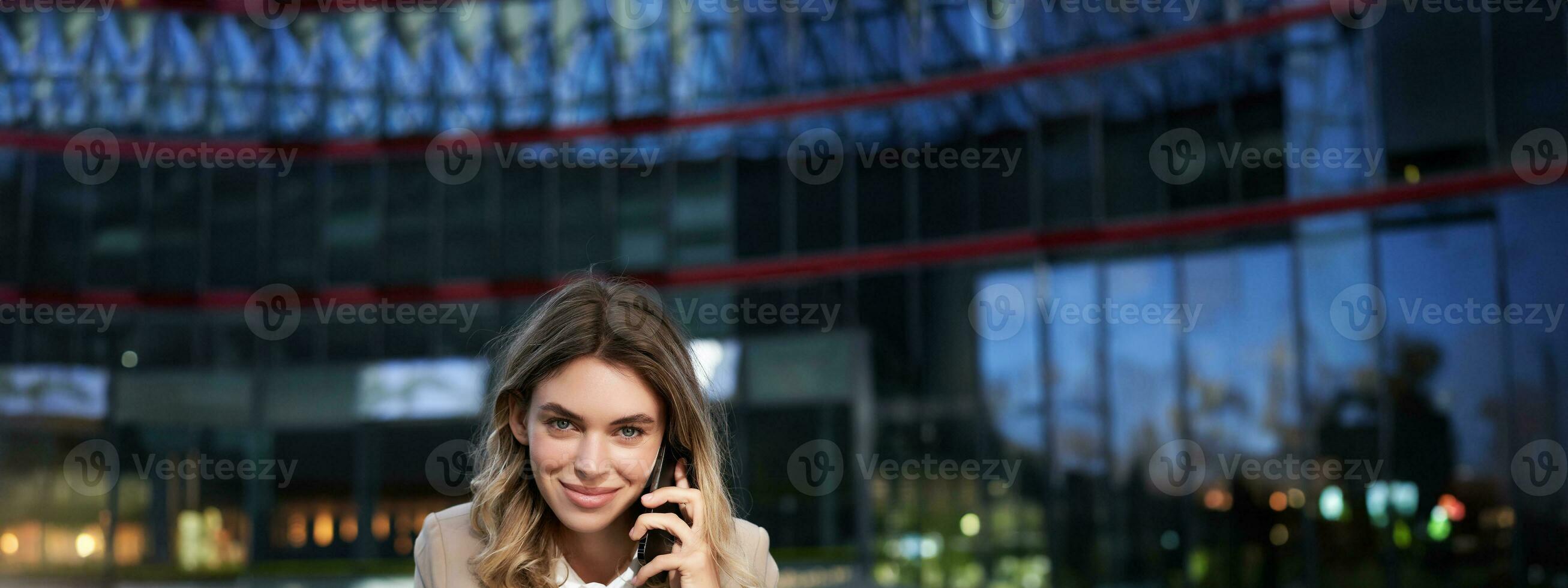 Vertical shot of smiling businesswoman answer mobile phone call, talking on smartphone and drinking coffee photo